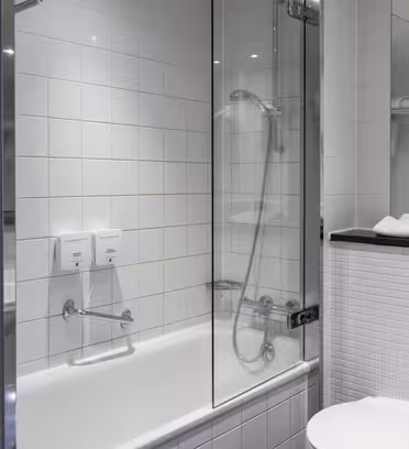 Moderne bad med dusj- og badekarkombinasjon med hvite fliser bak glassvegg. Veggmonterte dispensere er ved siden av dusjen. I forgrunnen er det hvitt toalett og vask. Håndklær er pent stablet på en hylle.