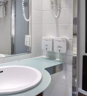 Moderne bad med hvit vask på benkeplate i glass, vegghengt hårføner, håndklestativ med oppbrettede hvite håndklær og stort speil som reflekterer motsatt vegg. Nøytralfargede fliser skaper et rent og organisert utseende.