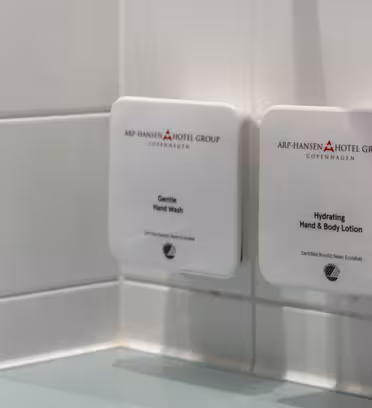 Nærbilde av to veggmonterte dispensere på bad. Den venstre dispenseren er merket "Gentle Hand Wash", og den høyre er merket "Hydrating Hand & Body Lotion." Begge er fra Arp-Hansen Hotel Group, København. Bakgrunnen har hvite fliser.
