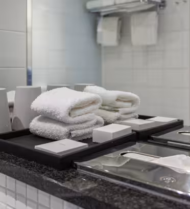Benkeplate for hotellbad med pent brettede hvite håndklær, hvite såpestykker på svarte brett og sølvpapirdispensere. Et speil reflekterer oppsettet, og det er en frostet glassvegg i bakgrunnen.
