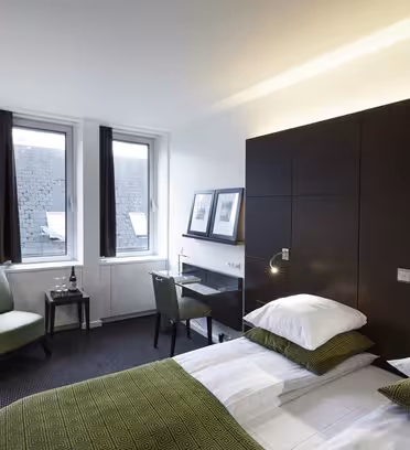 Et moderne hotellrom med en pent oppredd seng med grønne puter og dyne. Rommet har to store vinduer, et lite skrivebord med stol, en lenestol og innrammede bilder på veggen. Belysningen er myk og stemningsfull.