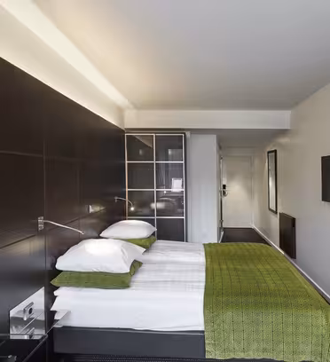 Moderne hotellrom med en pent laget dobbeltseng med grønt sengetøy. En flatskjerm-TV er montert på veggen nær et vindu. Rommet er dekorert med et enkelt innrammet bilde og har en minimalistisk design med mørke treaksenter.