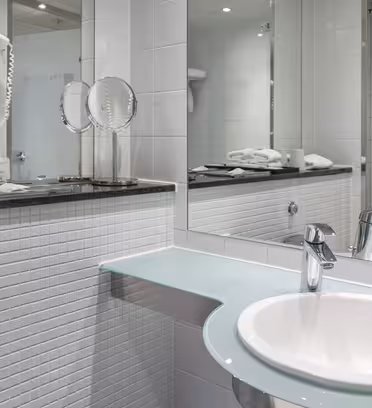 Et moderne bad med hvit vask og kromkran satt i benkeplate i glass. Et stort speil reflekterer scenen, og viser hvite flislagte vegger. Håndklær og toalettsaker er pent ordnet. Hårfønere er montert på veggen.