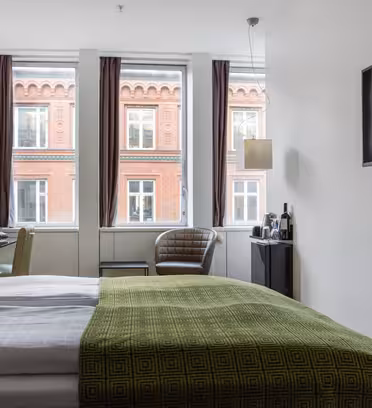 Et moderne hotellrom har en pent oppredd seng med grønt sengetøy, en veggmontert TV og store vinduer med utsikt over en rød mursteinsbygning med hvitkantede vinduer. Rommet inkluderer en stol, bord og hengende lamper.