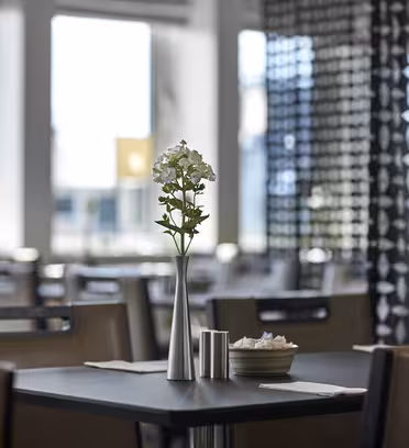 Et moderne restaurantinteriør med et bord med en sølvvase med hvite blomster. Bordet er dekket med en bolle og salt- og pepperbøsse. Myk belysning kommer gjennom store vinduer, og geometriske mønstre pryder en skillevegg.