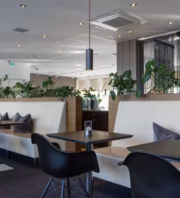 En moderne spisestue med svarte bord og stoler, beige polstrede boder og planter som pryder skilleveggene. Rommet har moderne belysning, store speil og et åpent takdesign, noe som skaper en elegant og innbydende atmosfære.