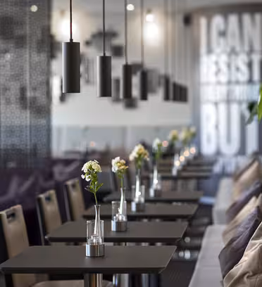 Moderne kaféinteriør med elegante svarte bord og stoler pyntet med små blomstervaser. Pendellamper henger over. Veggteksten sier "JEG KAN MOTSTÅ ALT MEN..." Stor plantekasse med grønt står mot en sofa med puter.