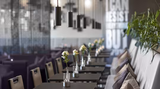 Moderne kaféinteriør med elegante svarte bord og stoler pyntet med små blomstervaser. Pendellamper henger over. Veggteksten sier "JEG KAN MOTSTÅ ALT MEN..." Stor plantekasse med grønt står mot en sofa med puter.