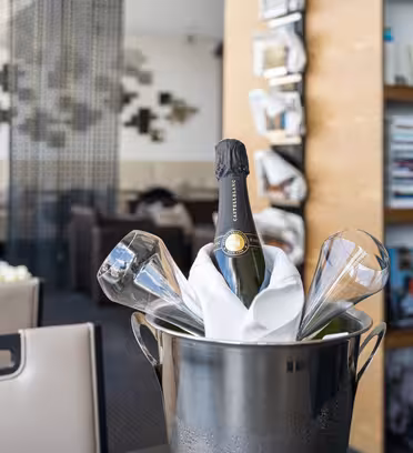 En flaske champagne avkjølt i en isbøtte med to glass står på et bord i en moderne, elegant spisestue. I bakgrunnen er uskarpe hyller med bøker og tidsriktig dekor synlige, noe som bidrar til en sofistikert atmosfære.